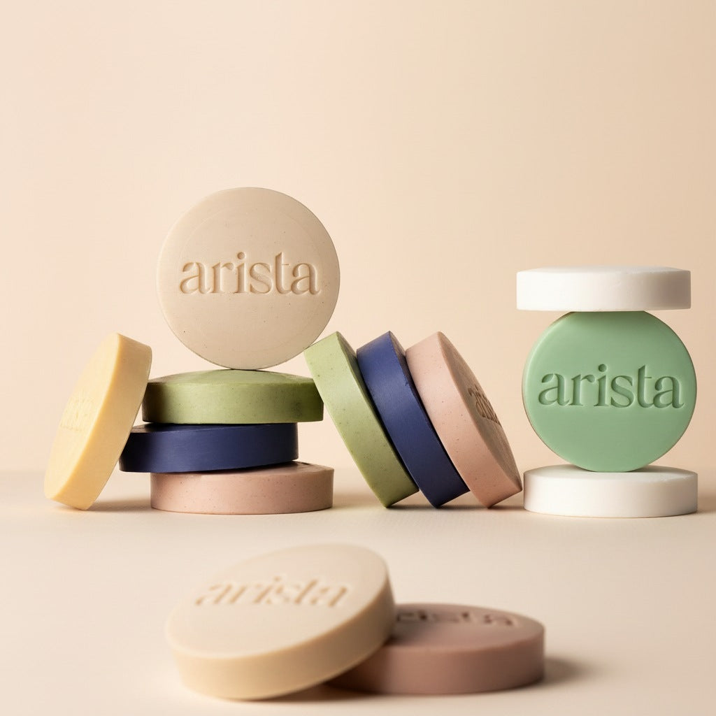 A set of round, disc-shaped shampoo bars branded 'arista' in embossed lettering, arranged on a light beige background. The bars come in soft pastel shades including cream, sage green, navy blue, dusty pink, and light yellow, with some stacked and some standing upright.