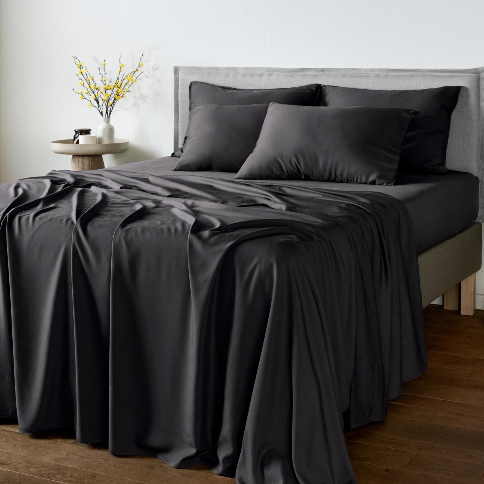 A neatly made bed with bambaw cozy black bamboo bedding, including a sheet, pillowcases, and a draped throw blanket, set in a modern bedroom with a gray upholstered headboard, wooden floor, and a small side table with yellow flowers.