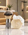 A bambaw metal shaving brush with dark and light vegan bristles and a textured silver handle stands beside a round cream-colored shaving soap bar with white lather on top, set on a marble countertop in a modern bathroom.